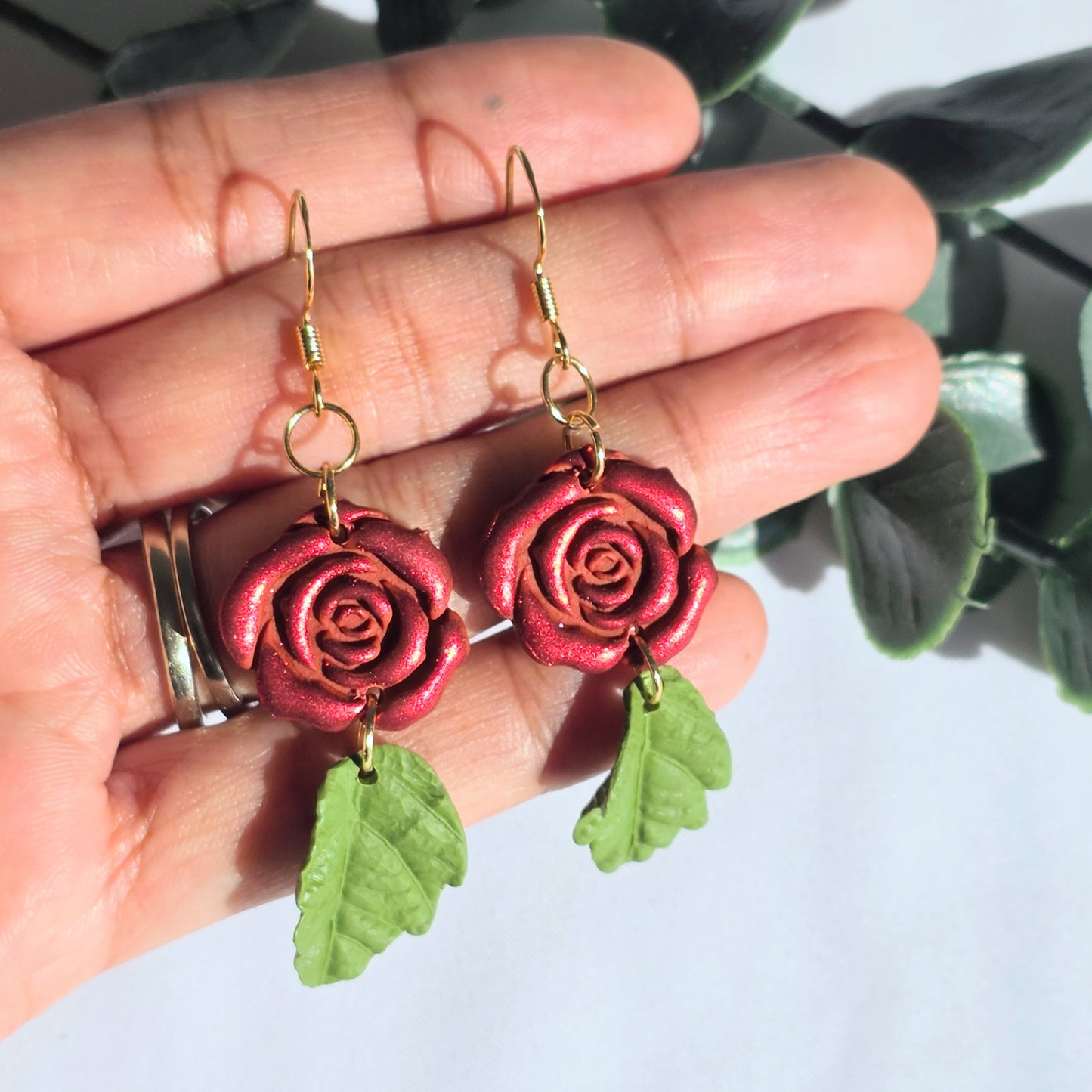 Rose-shaped earrings with green leaves held in a hand against a light background