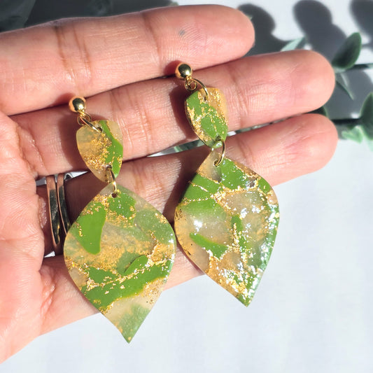 Green and gold leaf-shaped earrings held in a hand against a light background