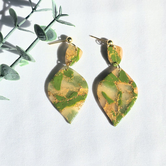 Green and gold leaf-shaped earrings on a white background with green leaves.