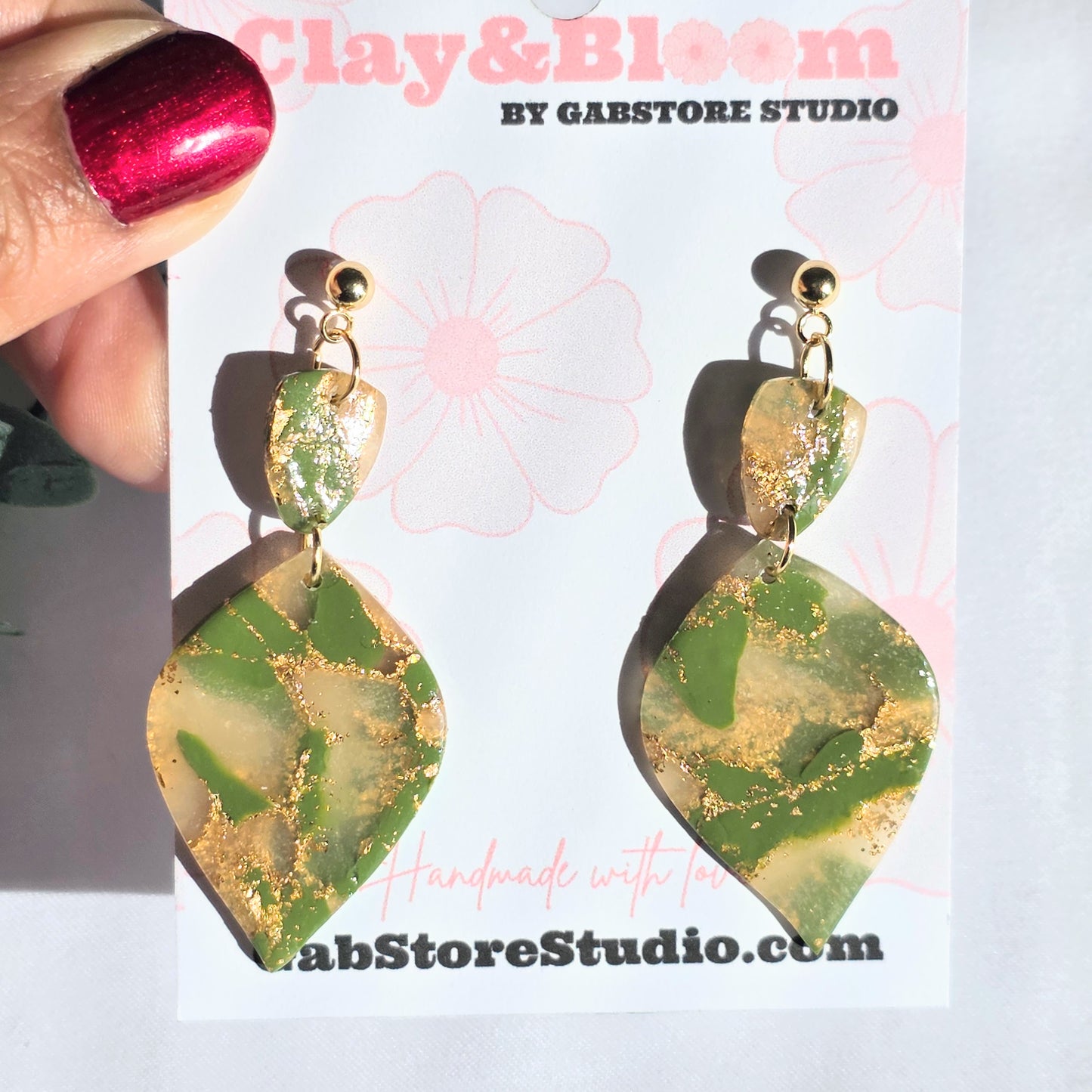 Green and gold earrings held by a hand with red nail polish, displayed on a white card with 'Clay & Bloom' branding.