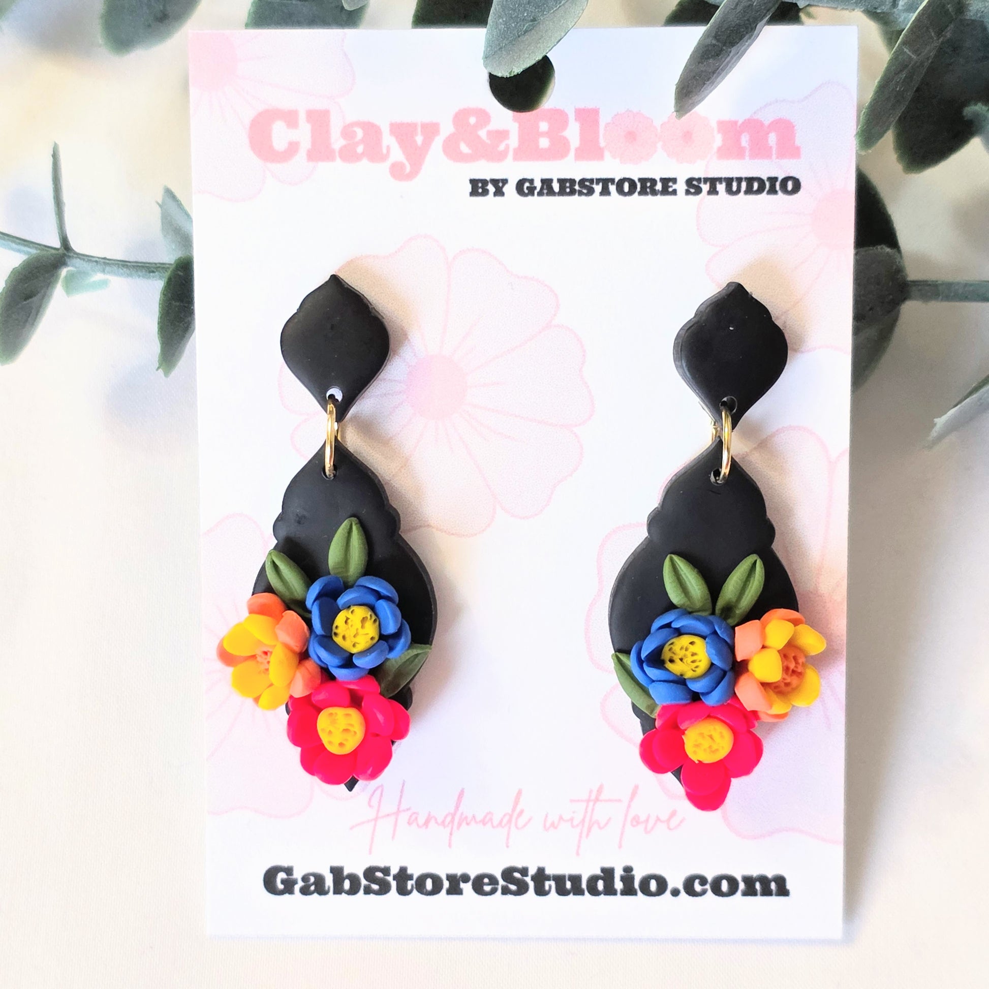 Black floral earrings on a 'Clay & Bloom by Gabstore Studio' card with green leaves in the background.