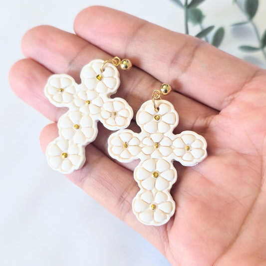 White cross-shaped earrings with gold accents held in a hand against a white background