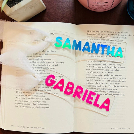 Open book with 'Samantha' and 'Gabriela' written on an acrylic bookmark, with a wooden surface and objects in the background.